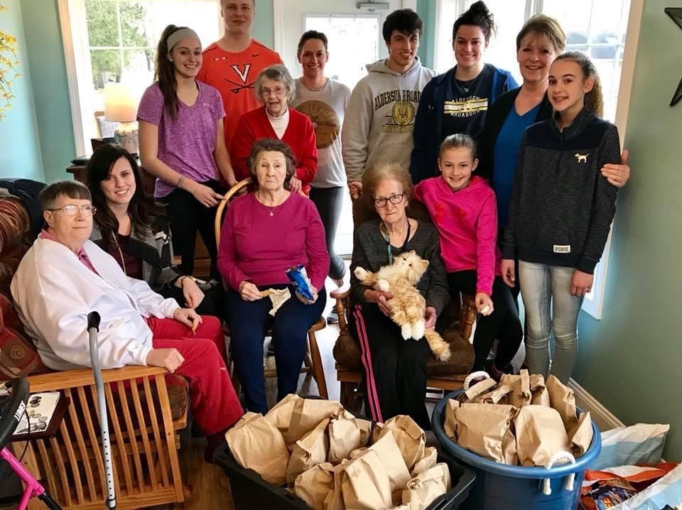 Group of people pose with bags. Indoors, smiling, diverse ages; woman holds cat, other person sits with walker.