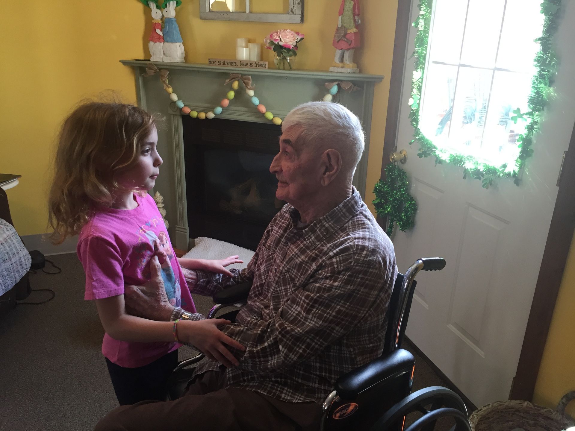 Girl in pink shirt talks to man in wheelchair in front of a fireplace.