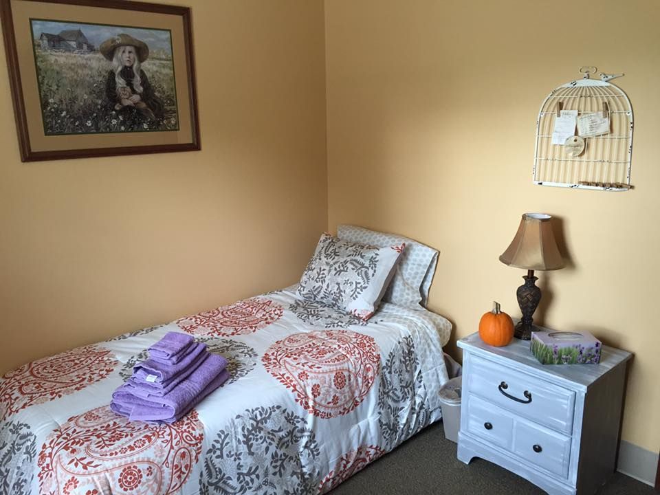 Small bedroom with single bed, nightstand, painting, and birdcage on wall. Orange and white bedding, purple towels.