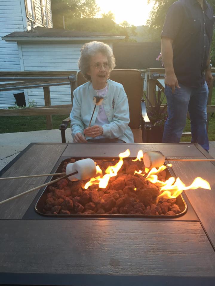 An elderly woman roasts a marshmallow over a fire pit, smiling outside.