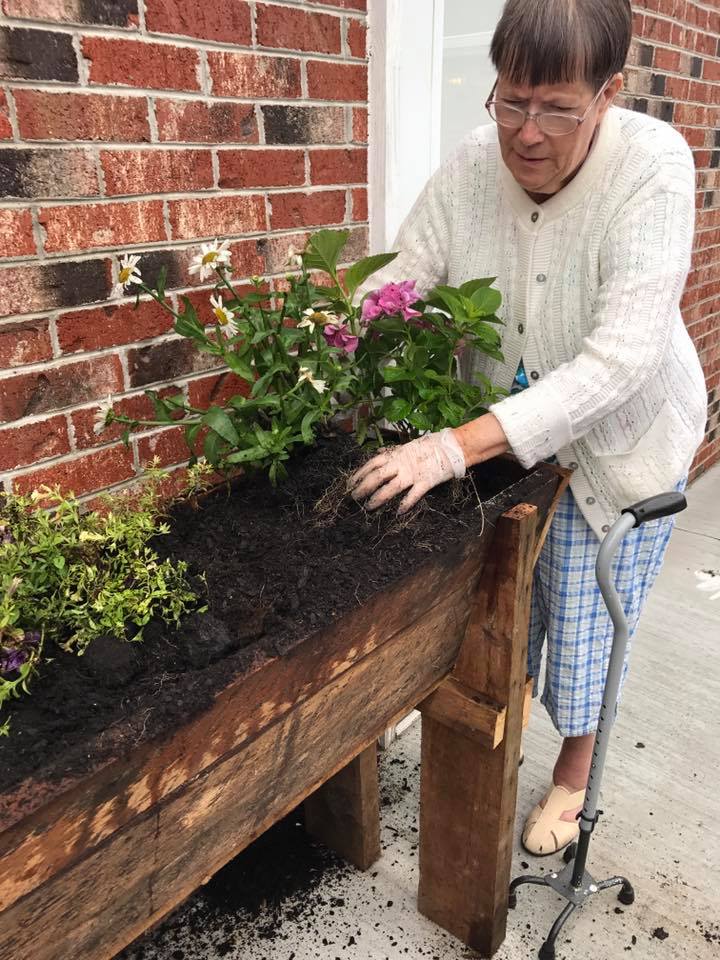 Woman in gloves gardening in a wooden planter box, using a cane for balance.