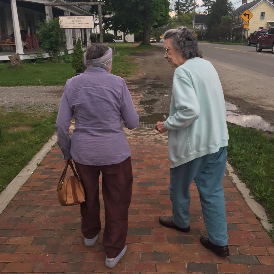 Two older women walking side-by-side on a brick sidewalk. One wears purple, the other light blue.