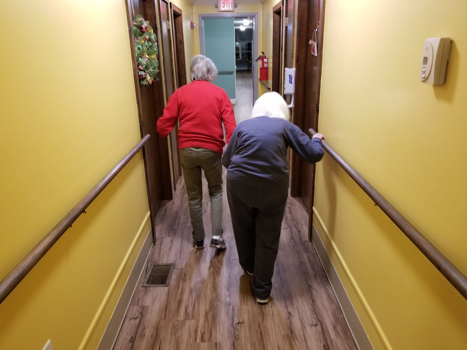 Two older people walk down a hallway, holding onto the wall railings. Yellow walls, brown floor, wooden doors.