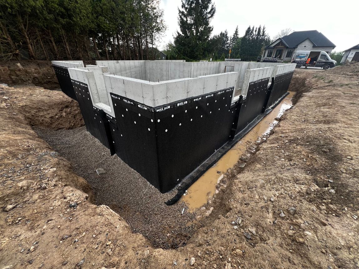 A large concrete foundation with waterproofing completed. The stone slinger has completed half of the backfill.