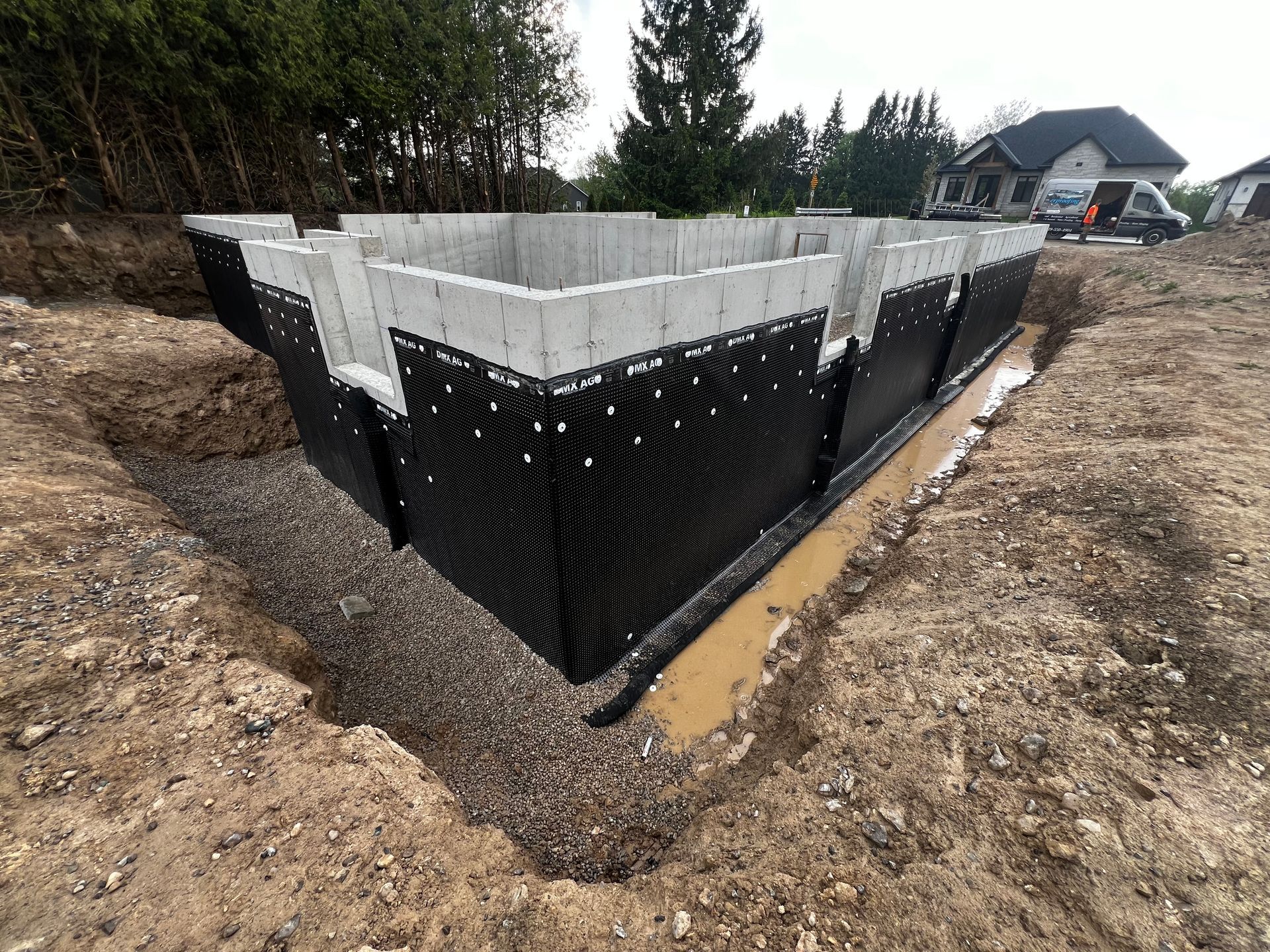 A large concrete foundation with waterproofing completed. The stone slinger has completed half of the backfill.