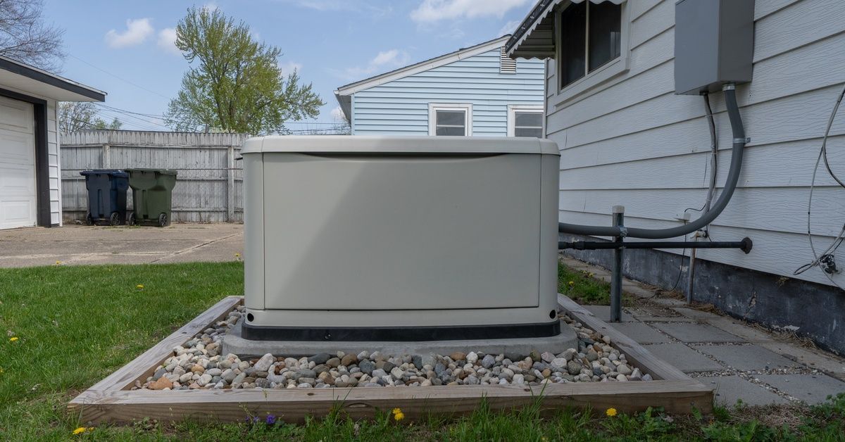 A gray home generator connected to a home with white siding. The generator sits on a bed of gravel i