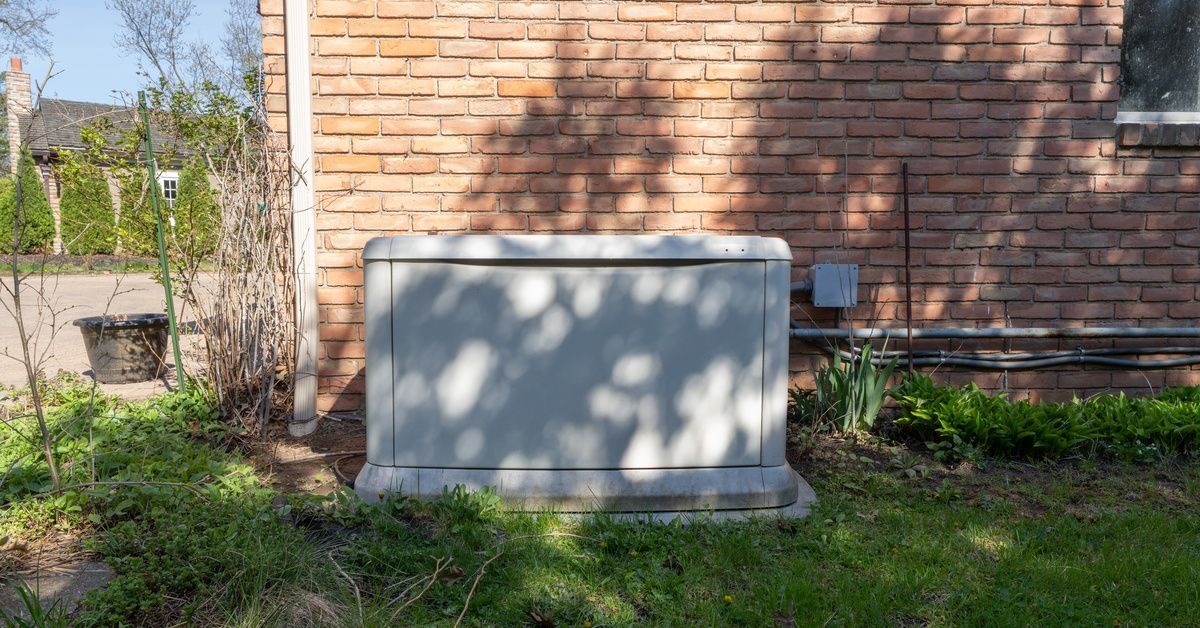 A power generator sits in front of a brick building. A tree is casting its shadow on the unit.