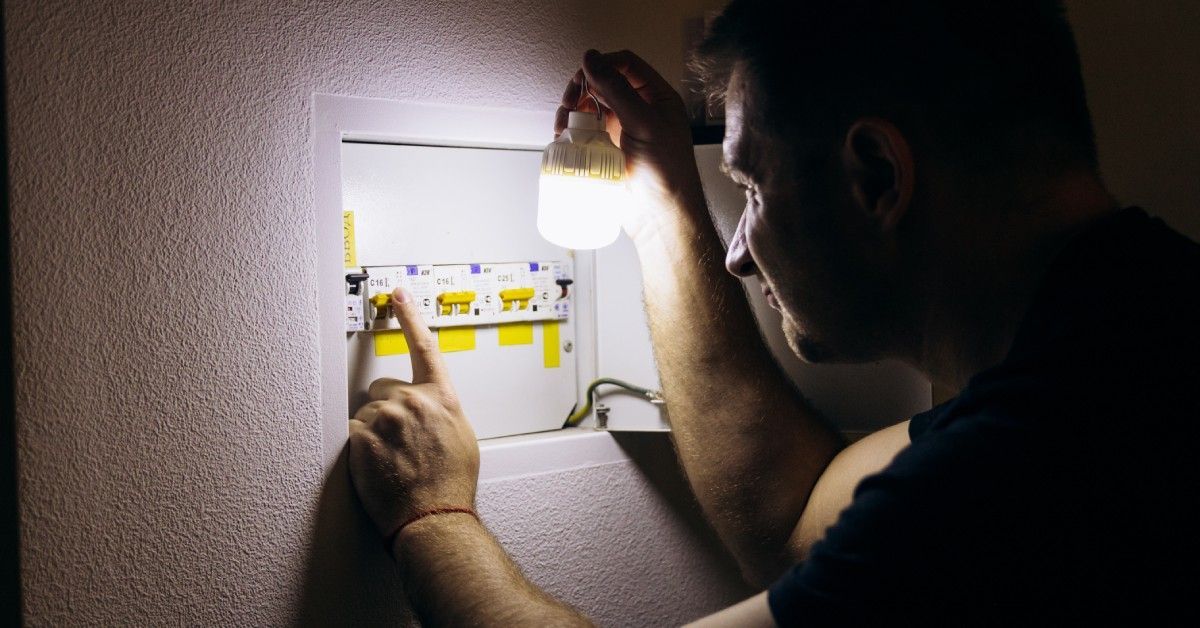 A person holds a bright light up to a breaker box in a pitch-black room. The person is squinting.