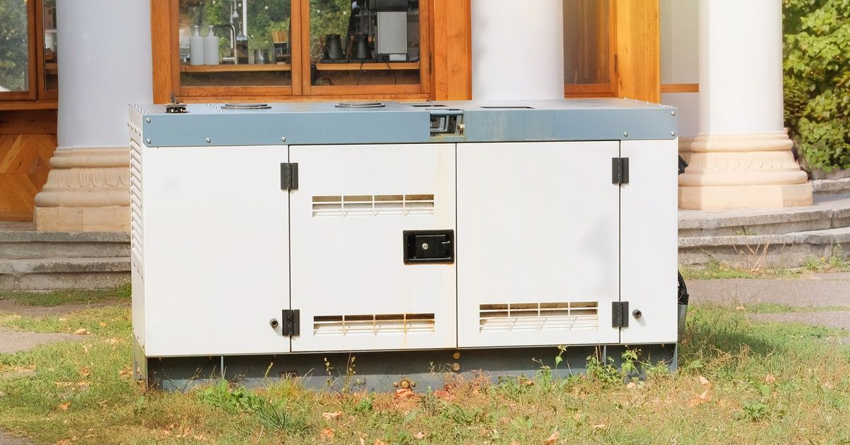 A diesel backup power generator sits in the grass outside of a building with wood-framed windows.