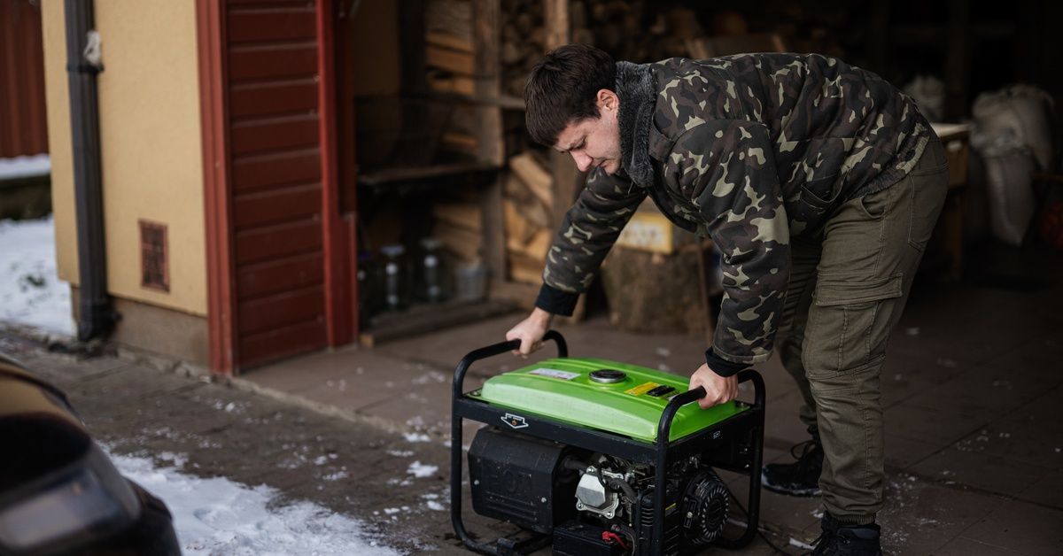 A man wearing a camouflage jacket is handling a portable gas backup generator outside of a garage.