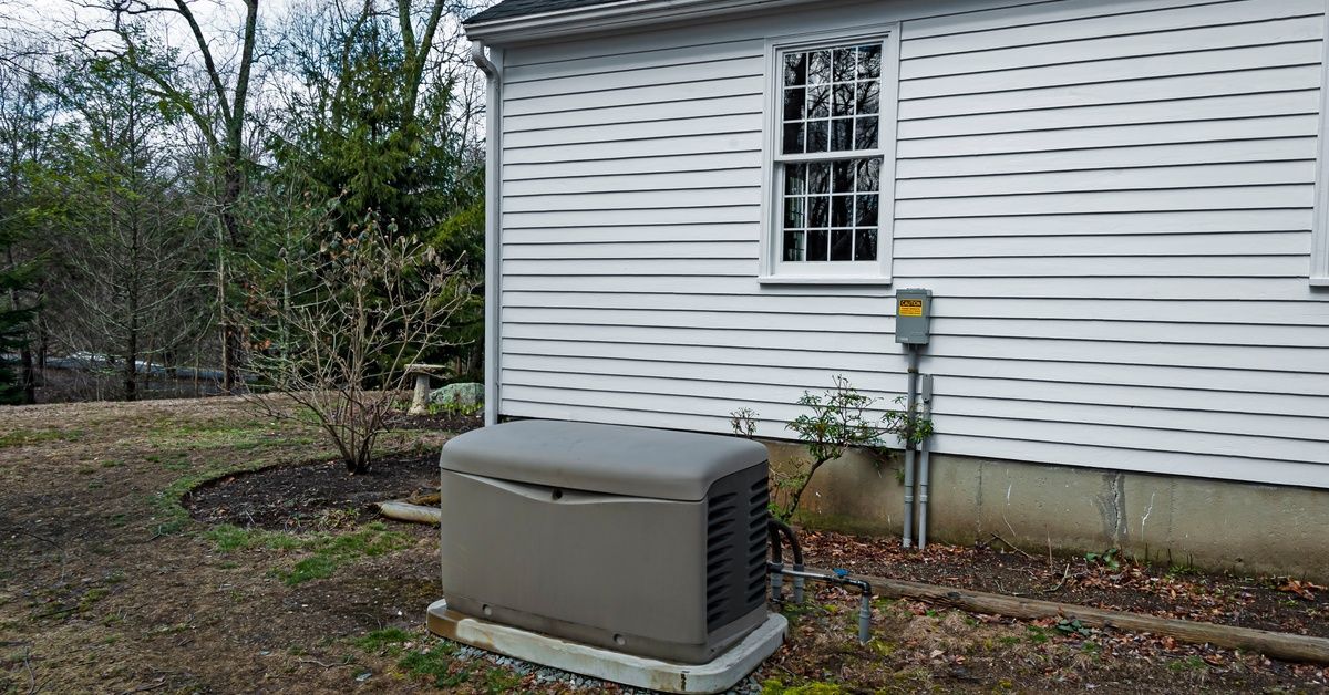 A gray residential power generator sits on a concrete slab on a lawn in front of a home with white s