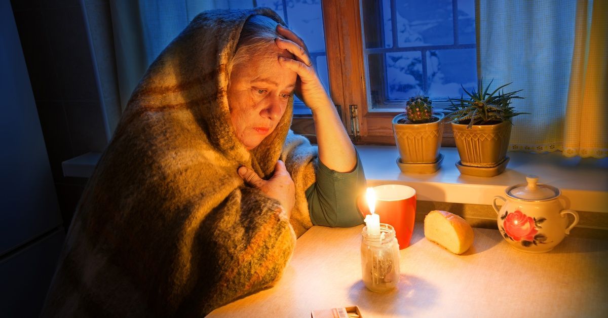 An elderly woman wrapped in a blanket sits in a dark room with a candle lit and a box of matches nex