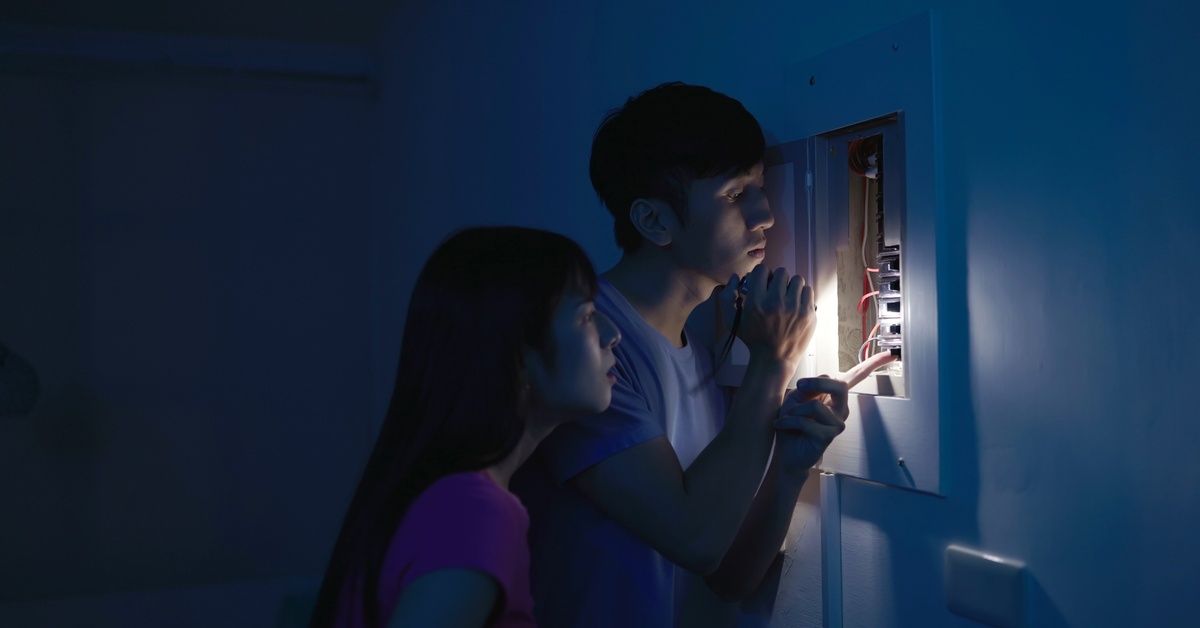 A man and woman with a flashlight are in a dark room looking at a breaker panel and touching one of 