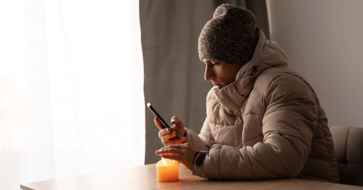 A man with a coat and hat is sitting at a table, putting his hand above a candle on the tabletop.