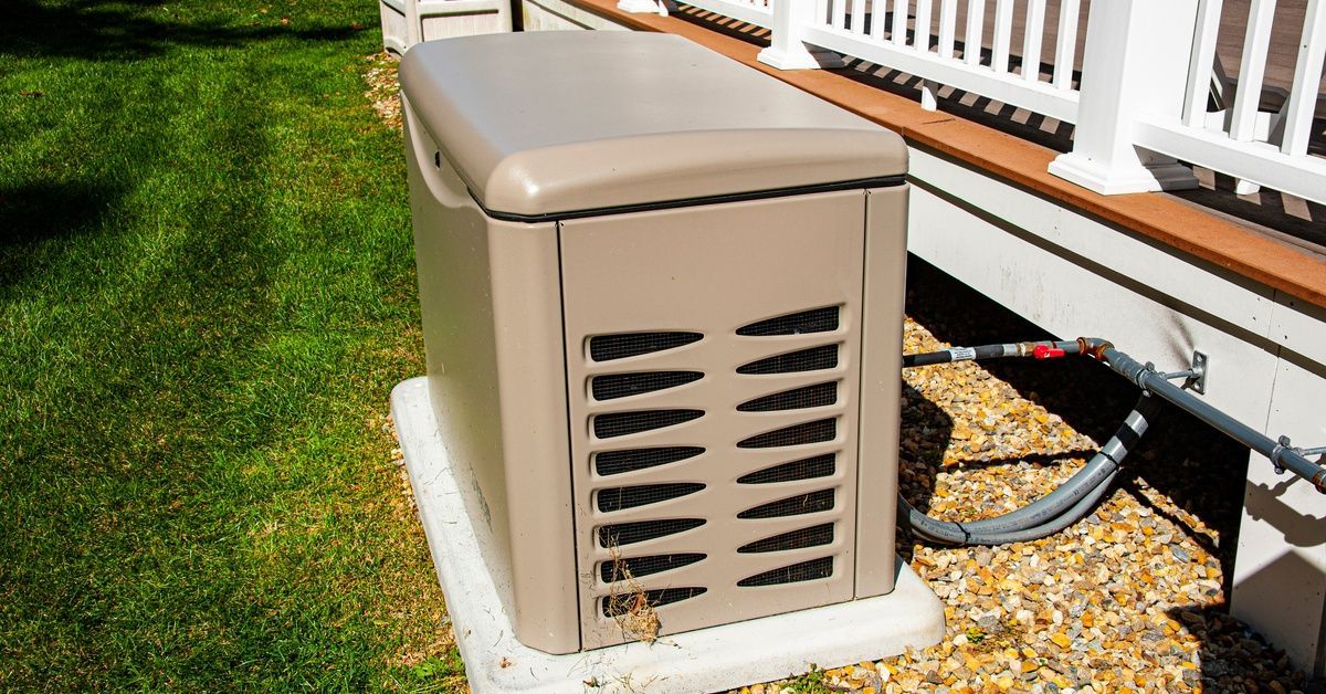 A brown generator on a concrete slab with pipes connecting to the side of a house in a grassy yard.