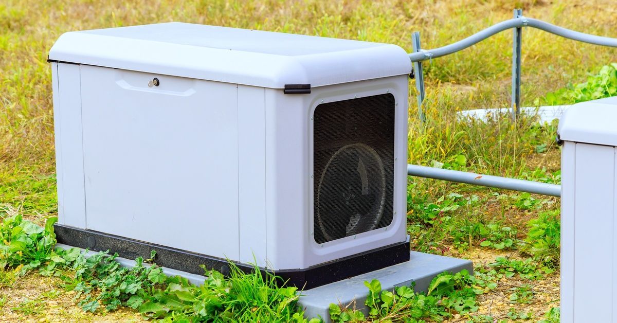 A gray generator with a visible fan sits on the grass next to some fencing and other equipment.