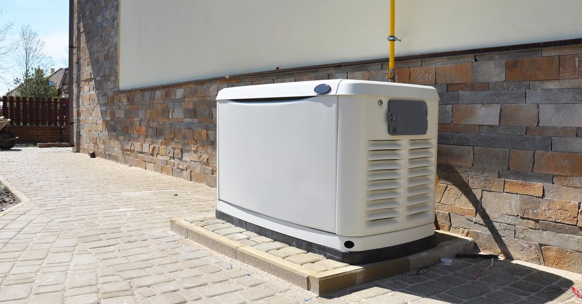 A house generator is alongside a brick house, sitting on a cement platform. A fence is nearby.