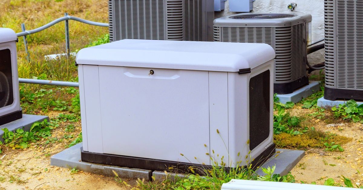 A home generator is sitting outside in a grassy and dirt yard, alongside multiple central air units.