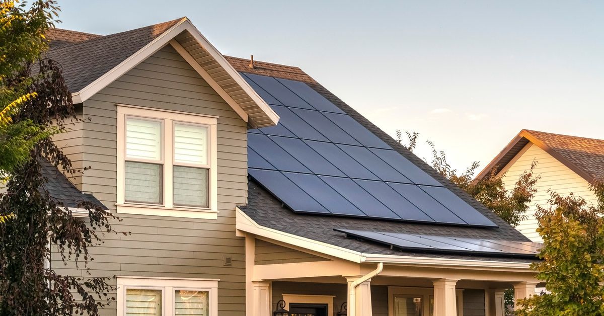 A two-story, grey-exterior home with solar photovoltaic panels installed on the roof on a sunny day.