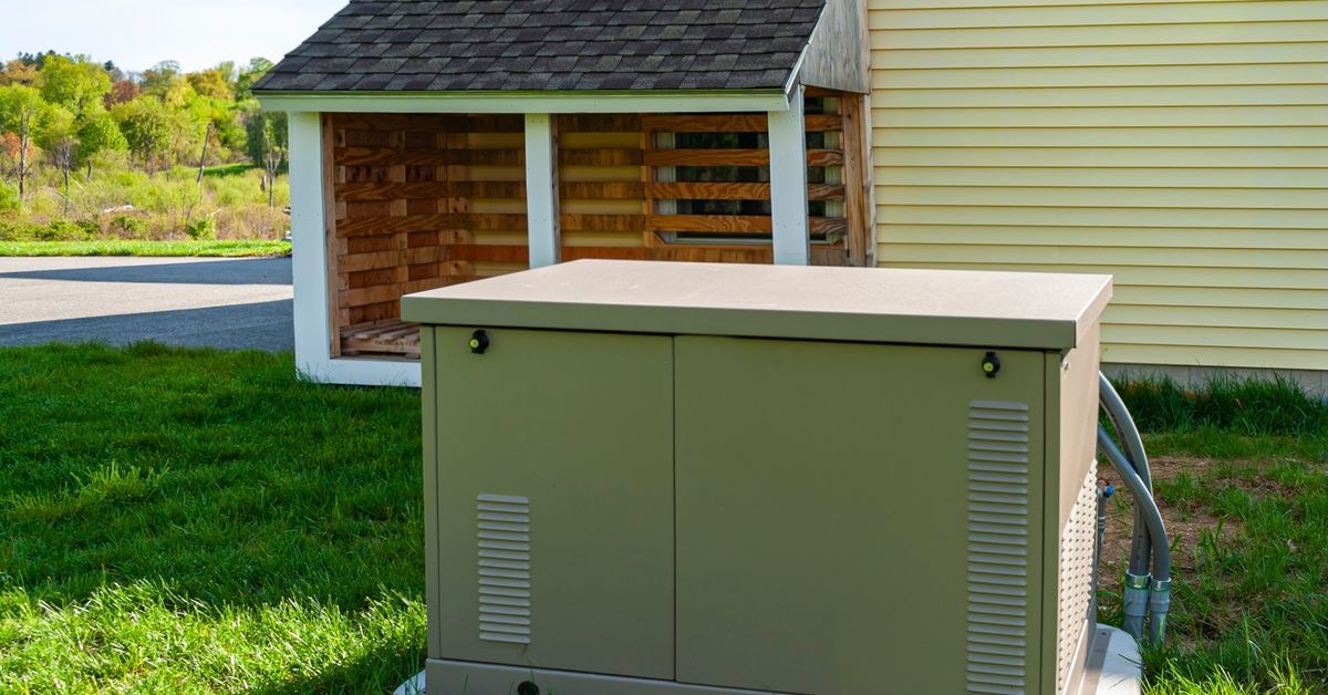 A dark-green home backup power generator sits on a lawn in front of a yellow home.