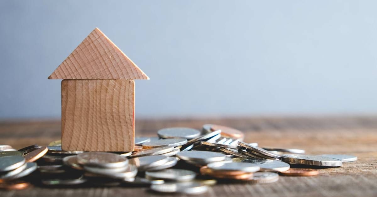 A home made of two wooden blocks sits on top of a pile of miscellaneous coins on a wooden table with