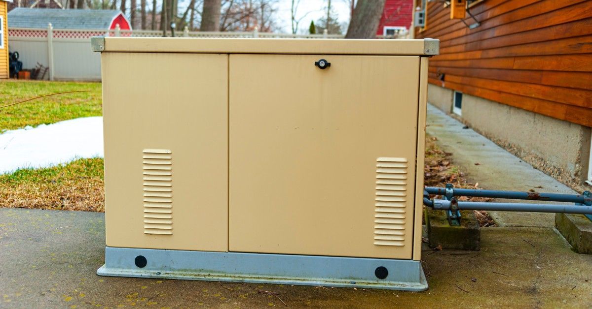 A tan, rectangular whole-home generator sits outside connected to a home with natural wood siding in