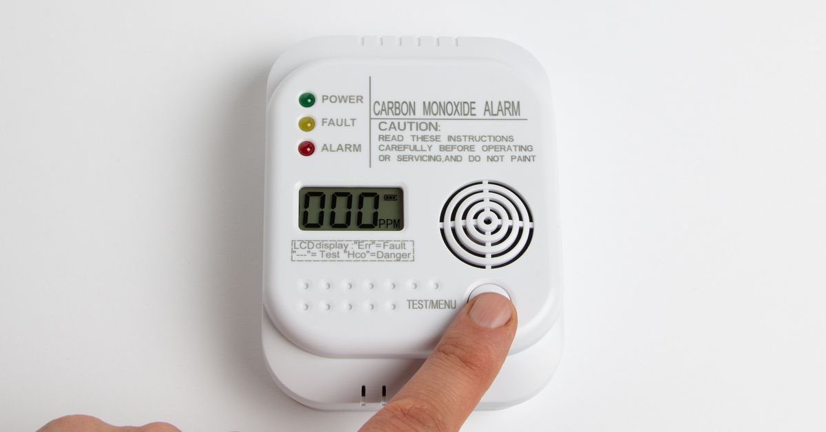 A rectangular carbon monoxide detector on a white wall. A person's finger presses a button on the bo