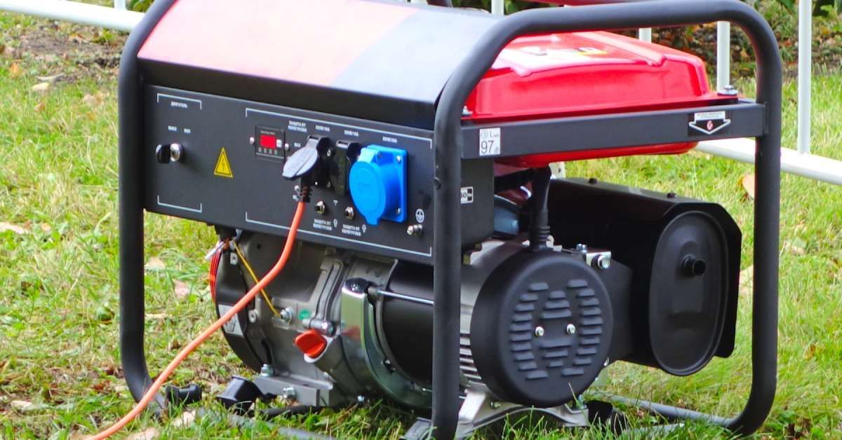 A compact, red portable generator with an orange cord on grass with a fence in the background.