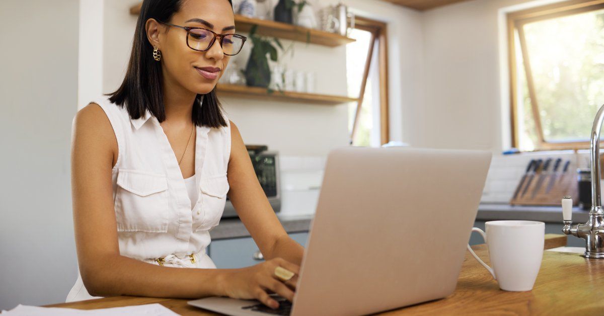 A woman wearing glasses and a white jumpsuit working on her laptop in her kitchen.