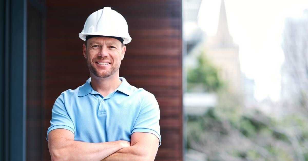 A contractor wearing a light blue polo and white hard hat stands smiling with his arms crossed.