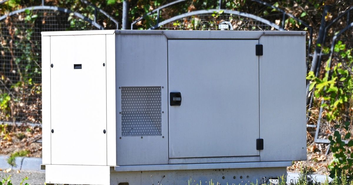 A generator installed in front of bushes enclosed by a fence. There is overgrowth in the distance.