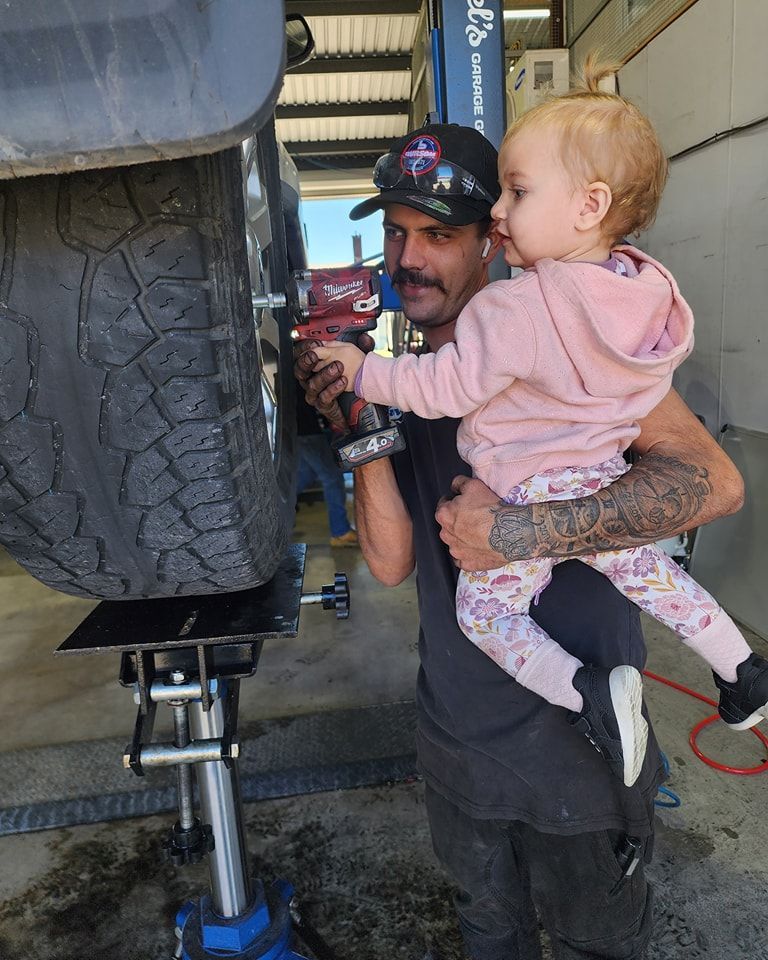 A Man Is Holding A Baby While Working On A Tyre — M8S Rates Mechanical In Gladstone Central, QLD