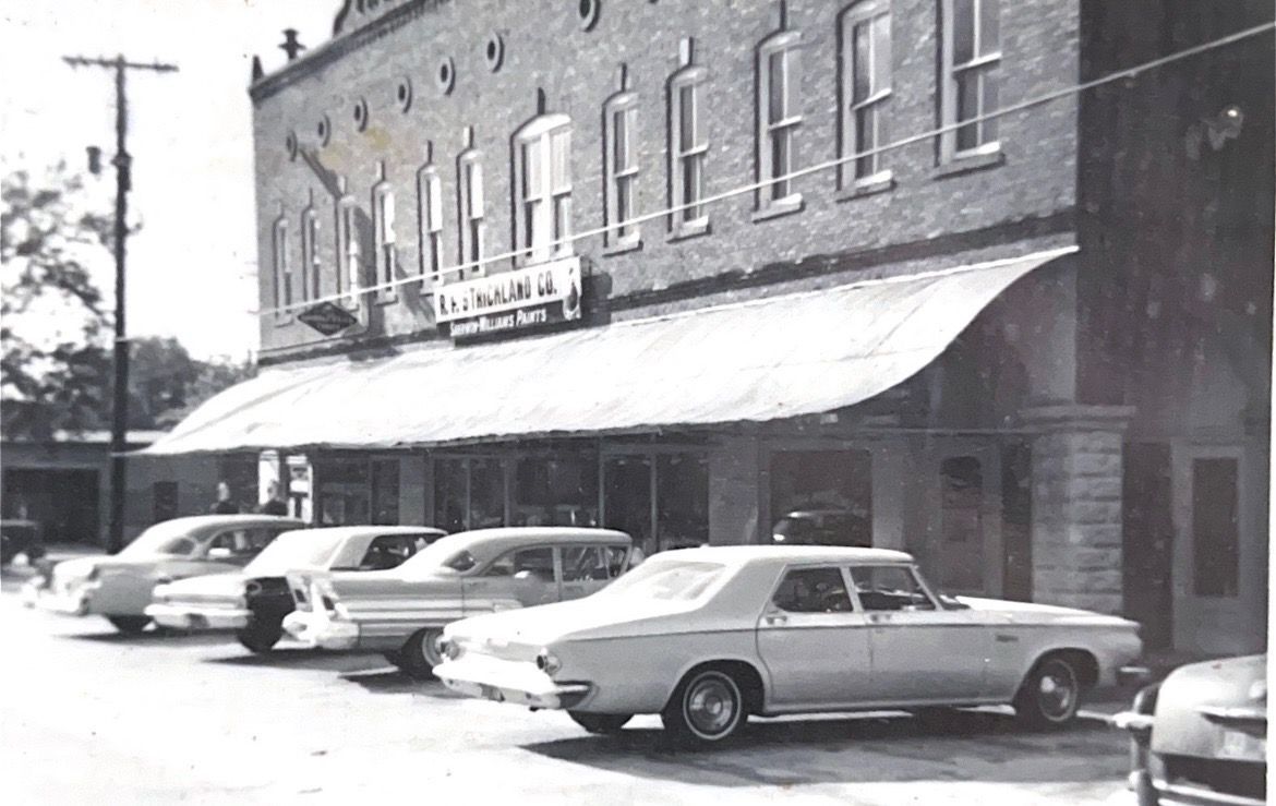 Black and white photo of a two-story brick building with a sign and cars parked out front.