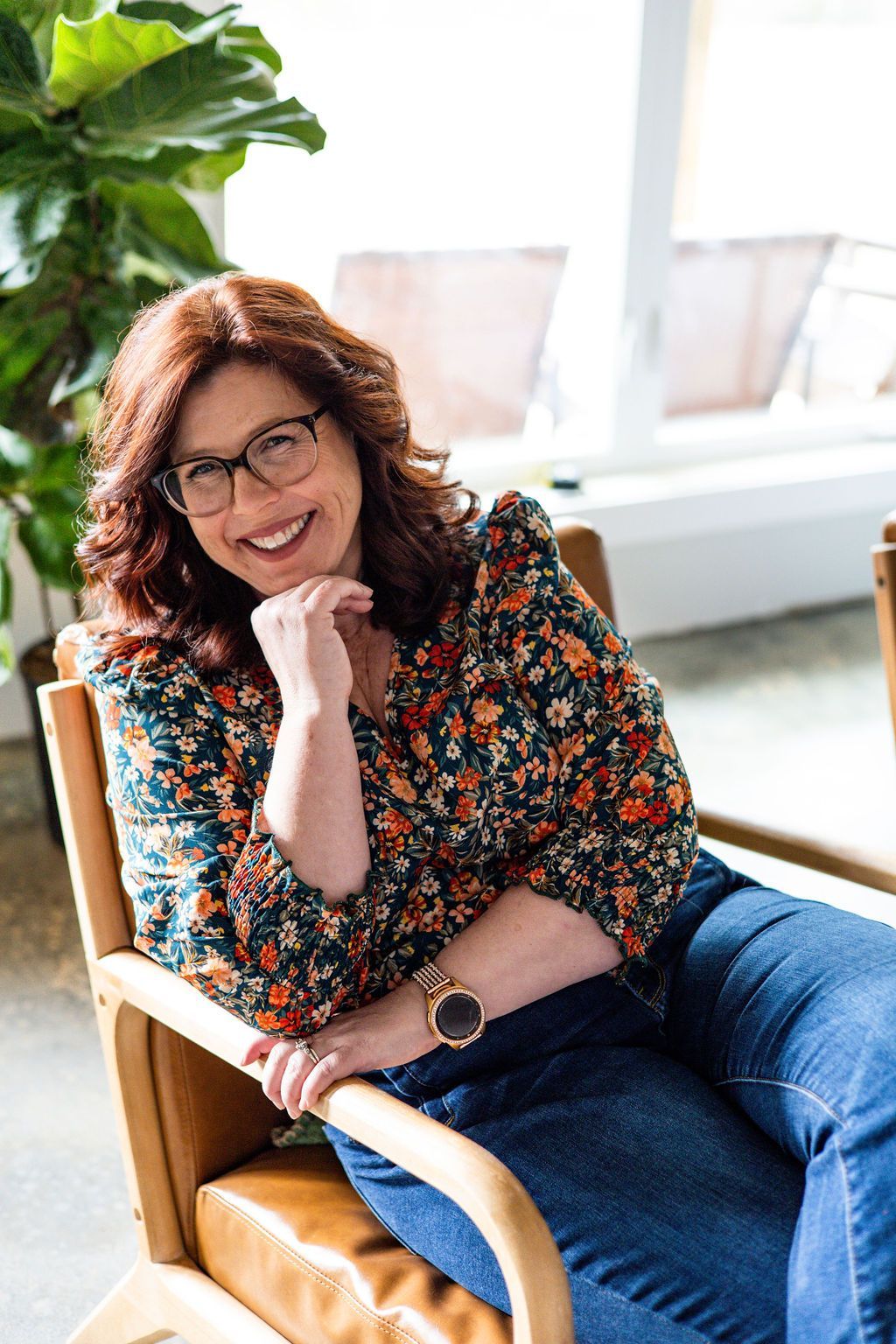 Woman in floral top smiles, leaning in chair, hand on chin.