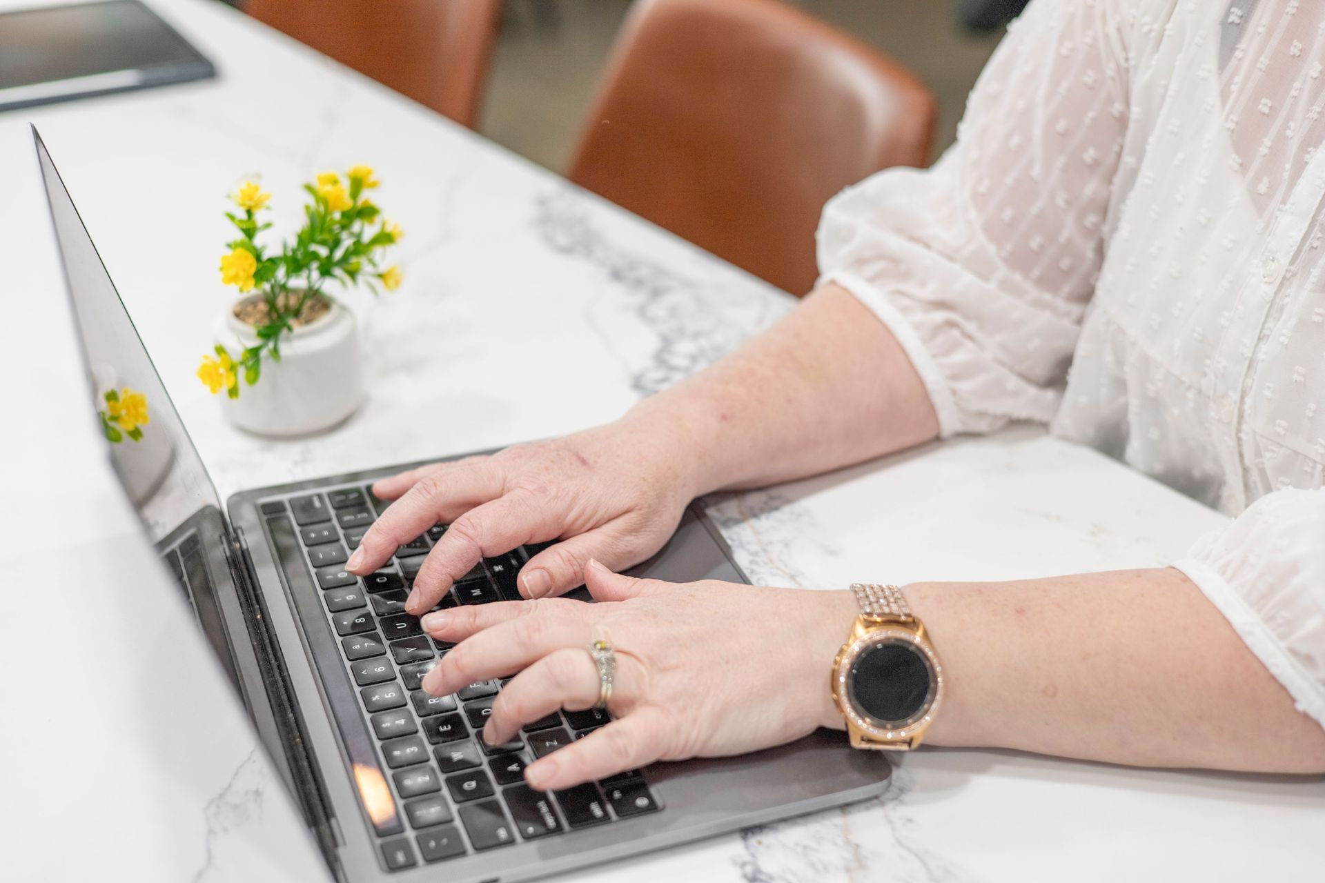 Person typing on a laptop at a marble table; small yellow flowers in a vase.