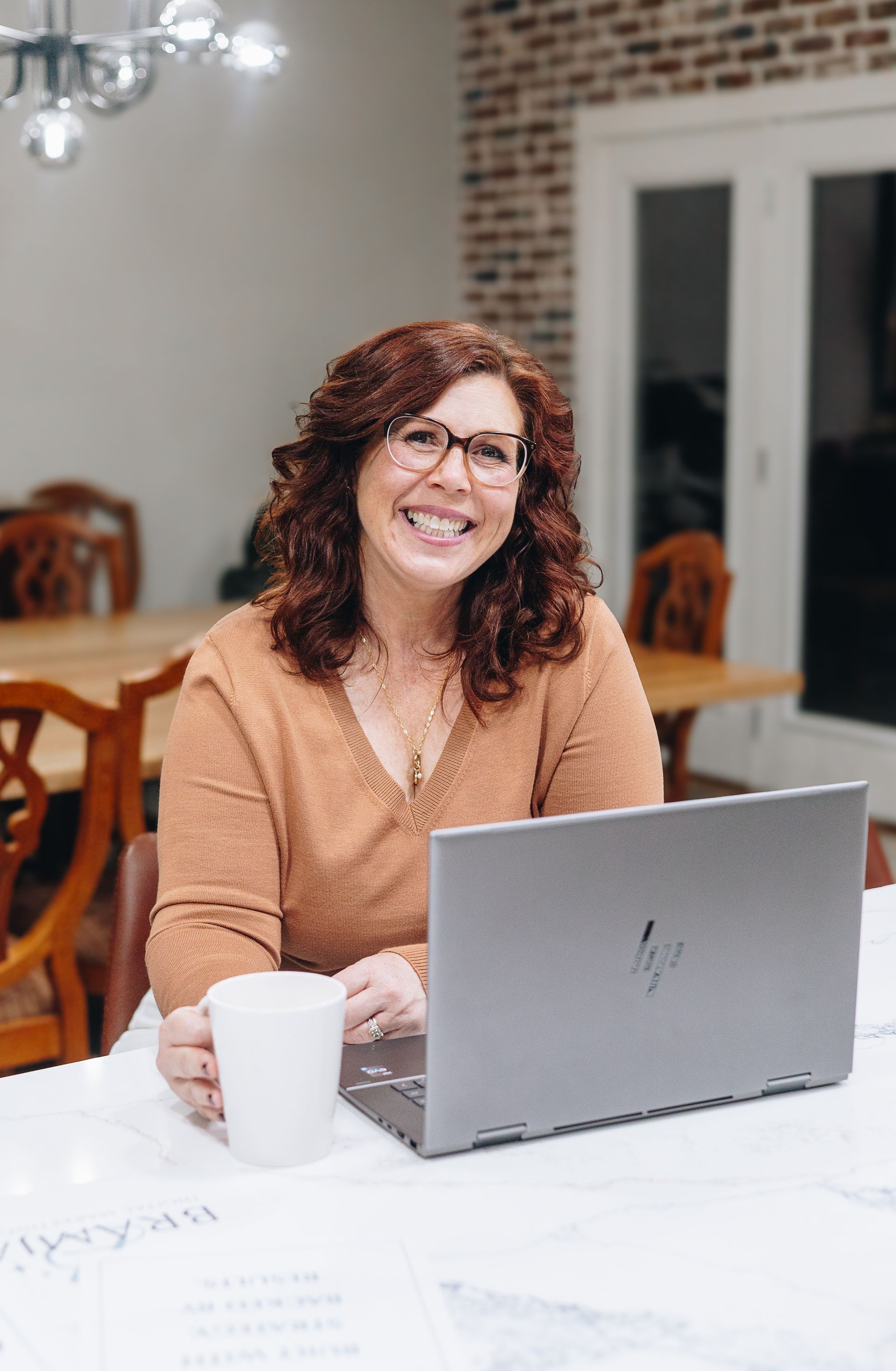 Woman wearing glasses, gray jacket, and jeans sits in a yellow armchair against an orange backdrop.