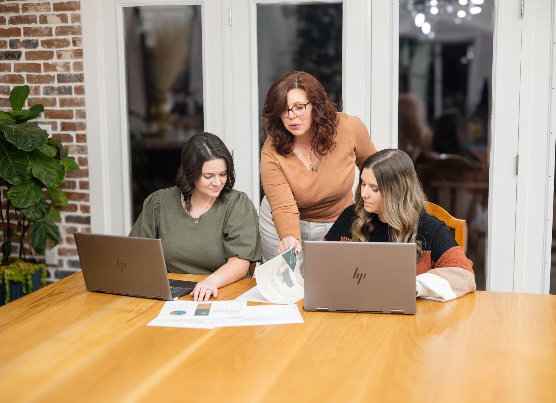 Three women reviewing documents together, working on laptops at a wooden table in a well-lit space.