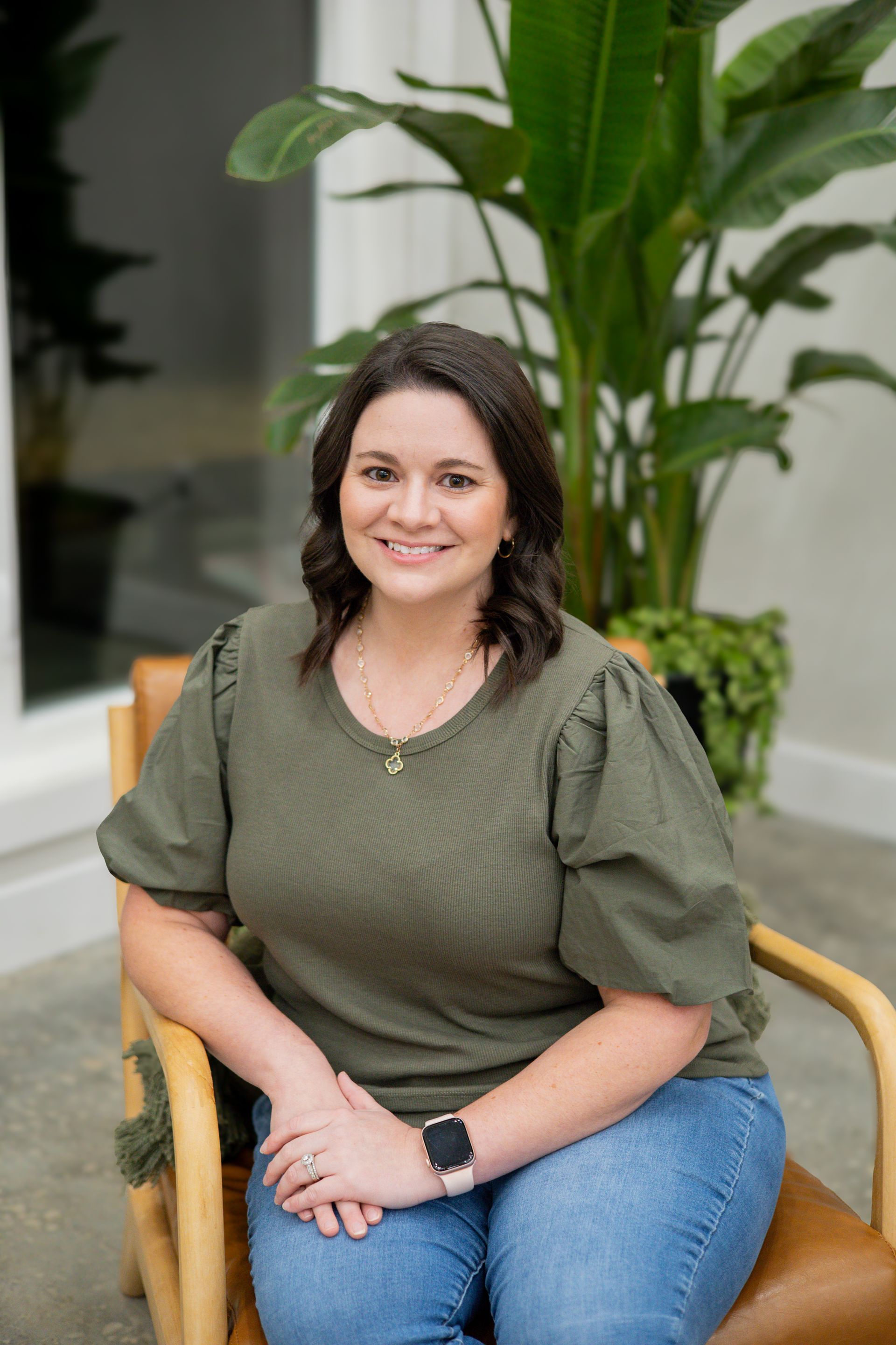 Woman in pink top and black pants smiling, seated on a pink chair, sunglasses on head.