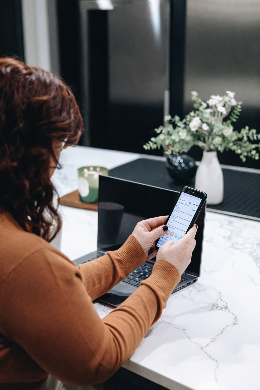 Woman using a smartphone and laptop at a white desk; silver pen. She wears a black top, ring, has dark nails.
