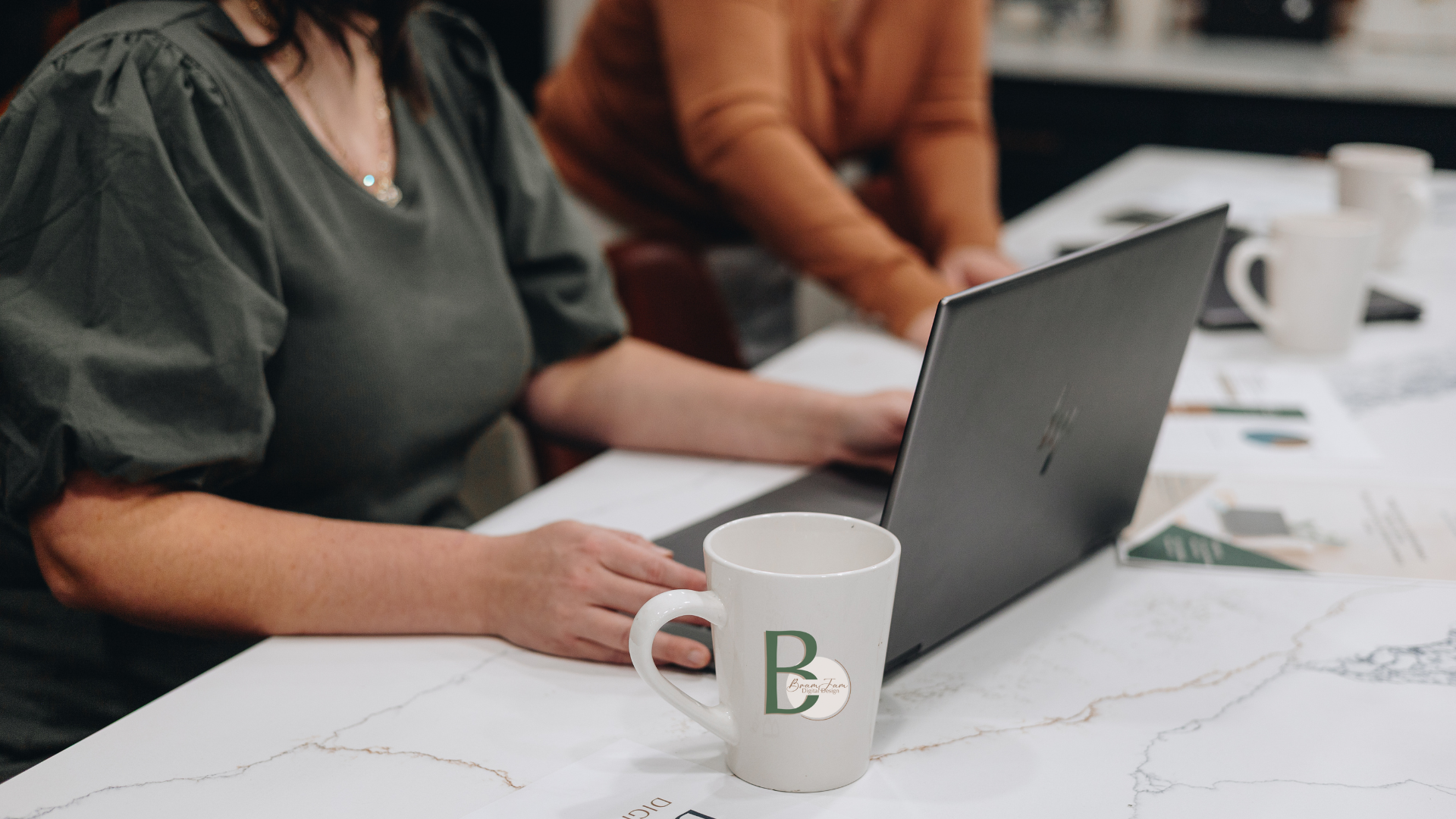 Person using a phone near a laptop and a drink tumbler on a marble countertop.