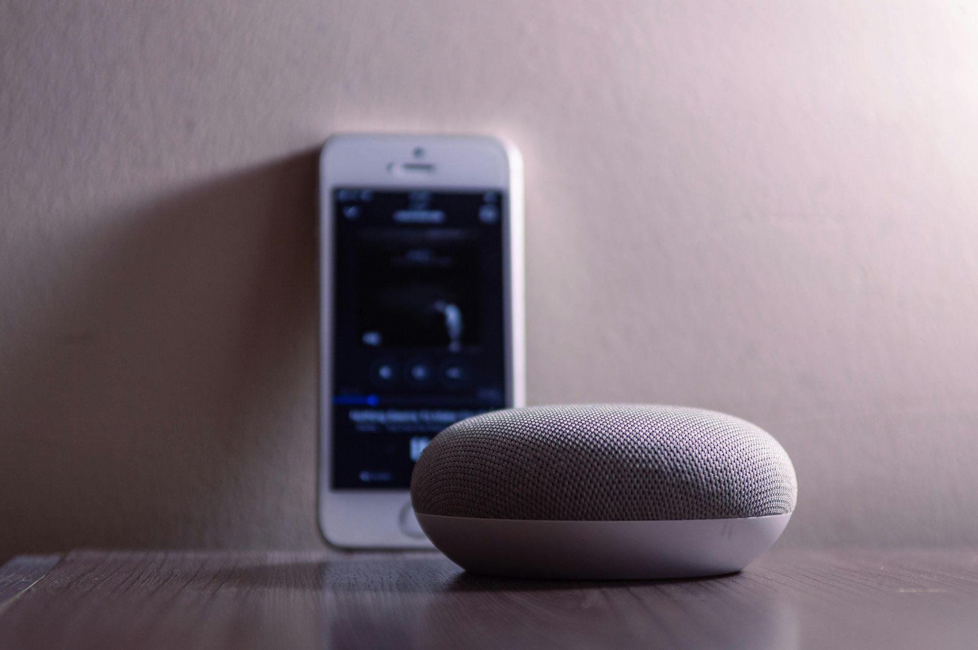 Small, gray smart speaker and smartphone on a wooden surface, against a light-colored wall.