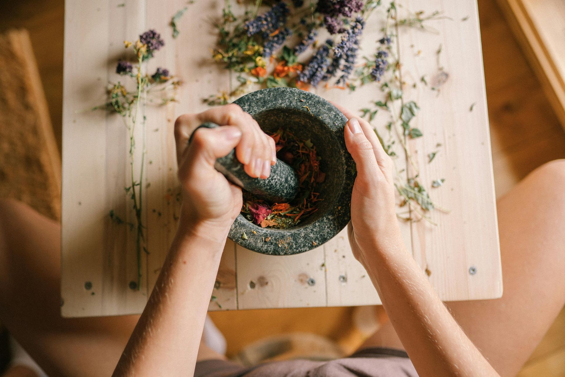 Person grinding herbs in a mortar and pestle on a wooden table, surrounded by more herbs.