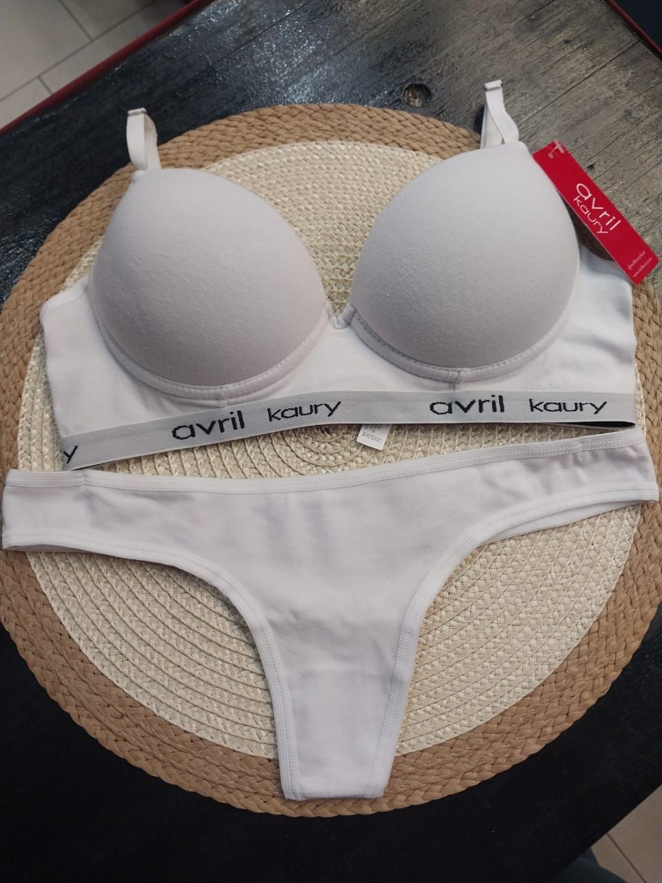 Conjunto de lencería blanca, sujetador con relleno, tanga, con nombre de marca sobre tapete tejido.