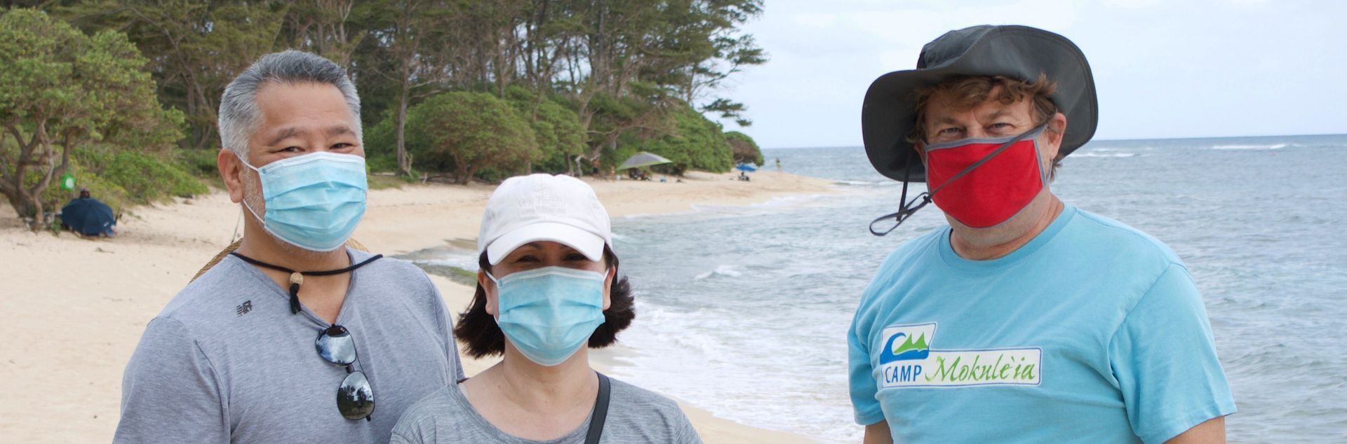Three people wearing face masks are standing on a beach.
