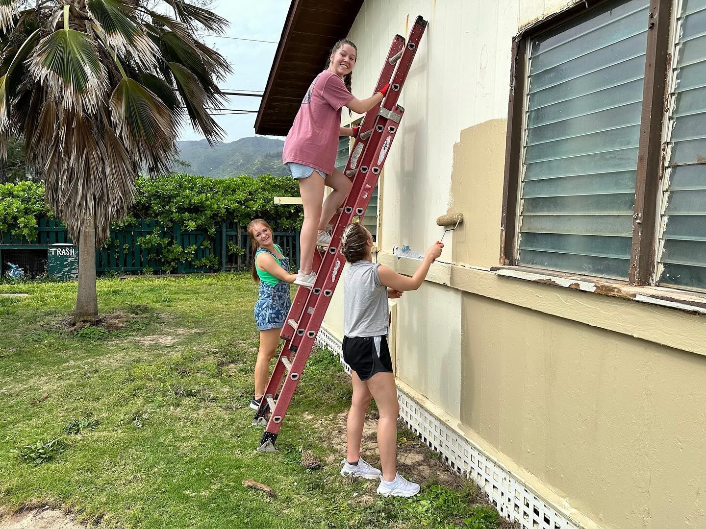 Three girls are painting a house with a ladder.