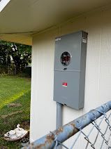 A electrical box is mounted on the side of a building next to a chain link fence.