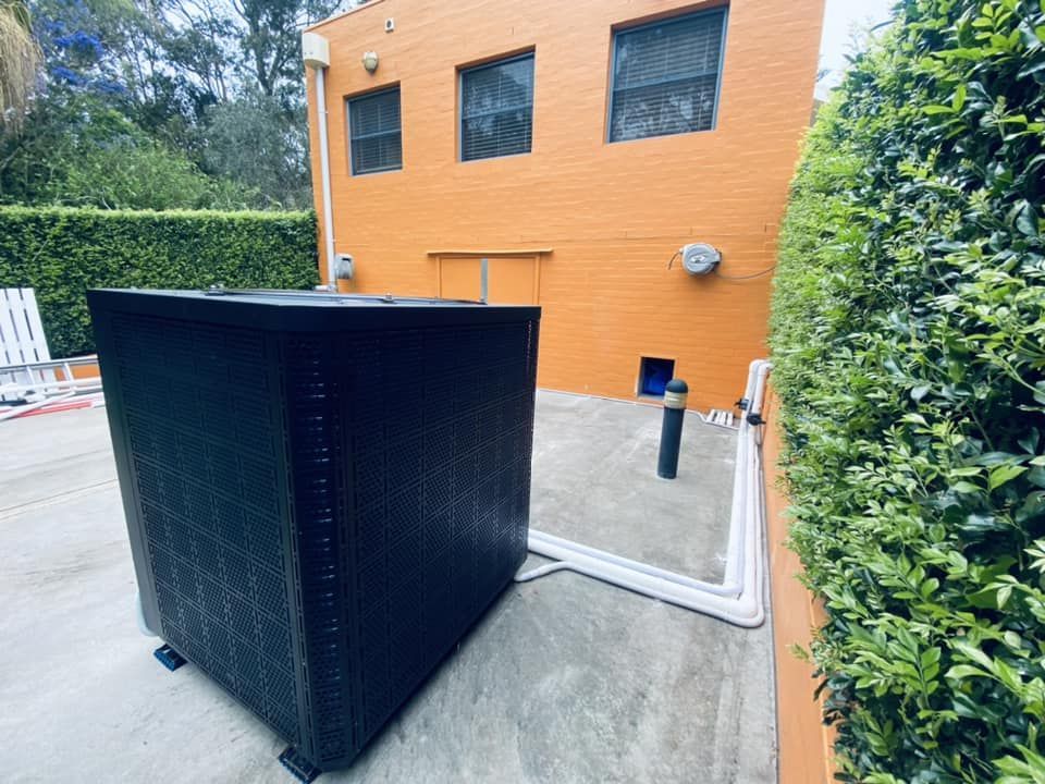 Black Pool Heater on a Concrete Surface Next to an Orange Building — Tempshift Pool Heating in Central Coast, NSW