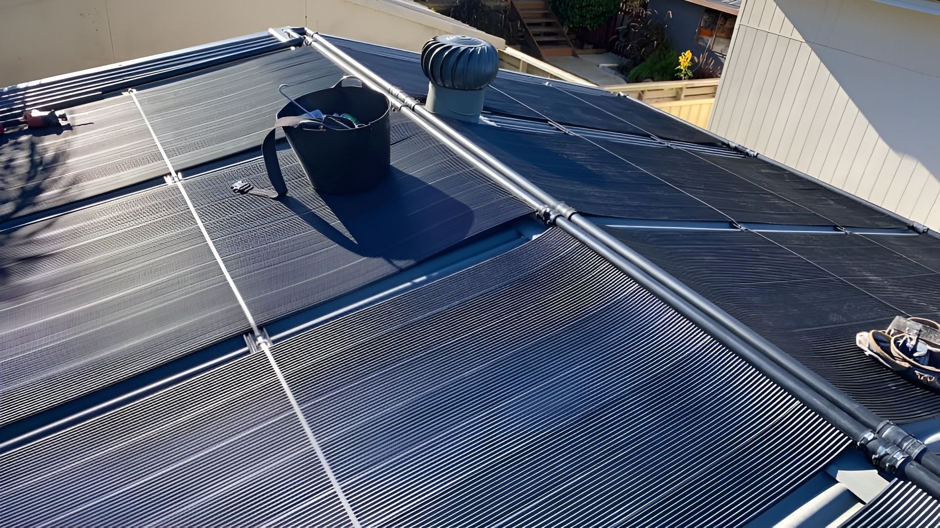 Black solar panels on a rooftop, with piping, a black bucket, and a rooftop vent — Tempshift Pool Heating in Buff Point, NSW