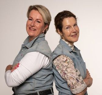 Twee lachende vrouwen staan met hun ruggen naar elkaar toe, gekleed in spijkerjasjes. Witte achtergrond.