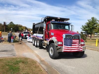 Red Mack dump truck draped in a large American flag at a roadside procession